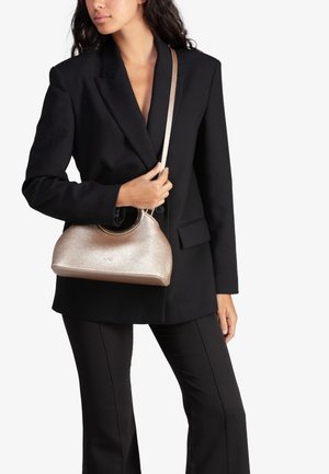 Gouden, metalen handtas met een ronde handgreep; gladde textuur; compact ontwerp, vastgehouden door een persoon in een zwarte blazer en broek.