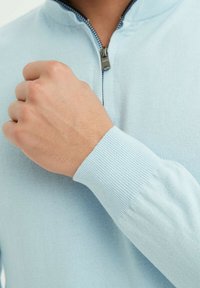 Pull à maille bleu clair avec un col zippé mi-hauteur, présentant des poignets côtelés et une texture lisse. Gros plan d'une main tenant le col.