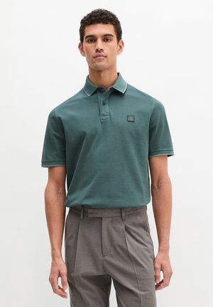 Mann trägt ein grünes Poloshirt mit kurzen Ärmeln und graue plissierte Hose, steht vor einem schlichten weißen Hintergrund.