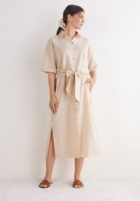 Beiges Leinenhemdkleid mit kurzen Ärmeln, Knöpfen vorne, einem gebundenen Taillengürtel, Seitentaschen und einem seitlichen Schlitz. Braune Sandalen.