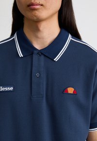 Mørkeblå polo-skjorte med hvite detaljer, med en pløse med to knapper og brodert logo på brystet med oransje og røde detaljer.