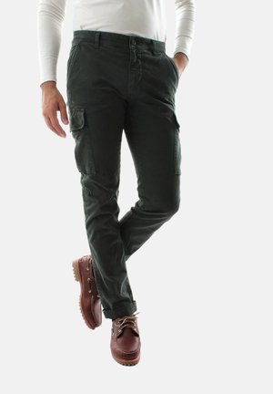 Persona che indossa pantaloni cargo verde scuro con tasche e orli arrotolati, scarpe di pelle marrone e una camicia bianca a maniche lunghe, in piedi con le gambe incrociate.