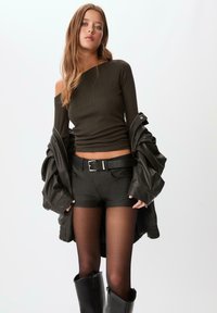 Sort læderjakke over en mørkegrøn ribbet off-shoulder top, parret med sorte shorts og knæhøje sorte støvler. Gennemsigtige sorte strømpebukser fremhæver benene.