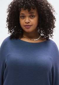 Σκούρο μπλε, πλεκτό τοπ με ριγέ υφή, ευρύ ντεκολτέ και άνετη εφαρμογή. Διαθέτει σγουρή υφή και συνδυάζεται με περιδέραιο από μαργαριτάρια και χρυσή αλυσίδα.