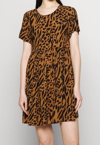 Kvinna som bär klänning med kort ärm och ovanför knät i brun och svart abstrakt leopardmönster mot en enfärgad bakgrund.