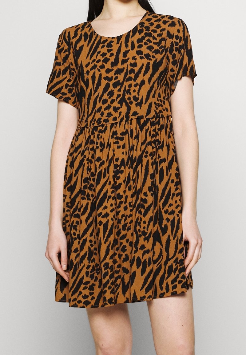 Kvinna som bär klänning med kort ärm och ovanför knät i brun och svart abstrakt leopardmönster mot en enfärgad bakgrund.