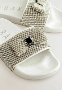 Baltas slaidu sandales ar teksturētu augšdaļu, rotātu ar mazām sudraba rhinestones un dekoratīvu loku, piestiprinātu ar melnu akcentu.