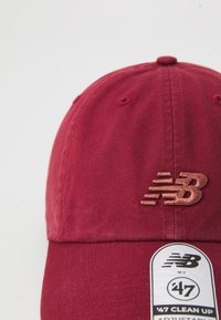Red cotton baseball cap featuring an embossed "NB" logo in copper detail. Adjustable strap with a hanging tag for size information.
