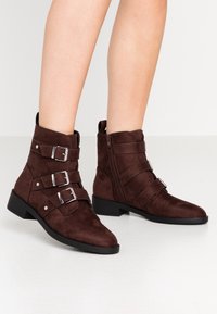 Botines de ante marrón con tres hebillas decorativas, cremallera lateral y tacón bajo, con un acabado texturizado y punta afilada.