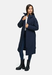 Manteau long bleu marine en tissu lisse, avec une capuche, fermeture éclair, poches latérales et coupe décontractée, associé à un jean foncé et des bottes noires.