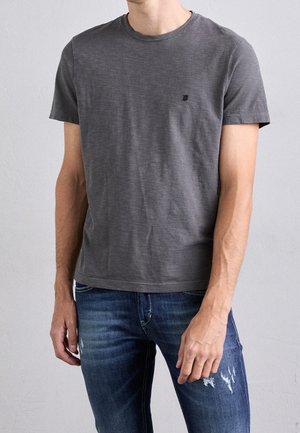 Homme portant un t-shirt à manches courtes gris foncé uni et un jean bleu délavé, debout devant un fond clair uni.