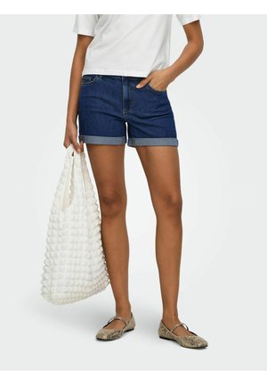 Femme portant un short en jean foncé retroussé, un t-shirt court blanc, des ballerines à imprimé serpent, tenant un sac fourre-tout blanc texturé à ses côtés.