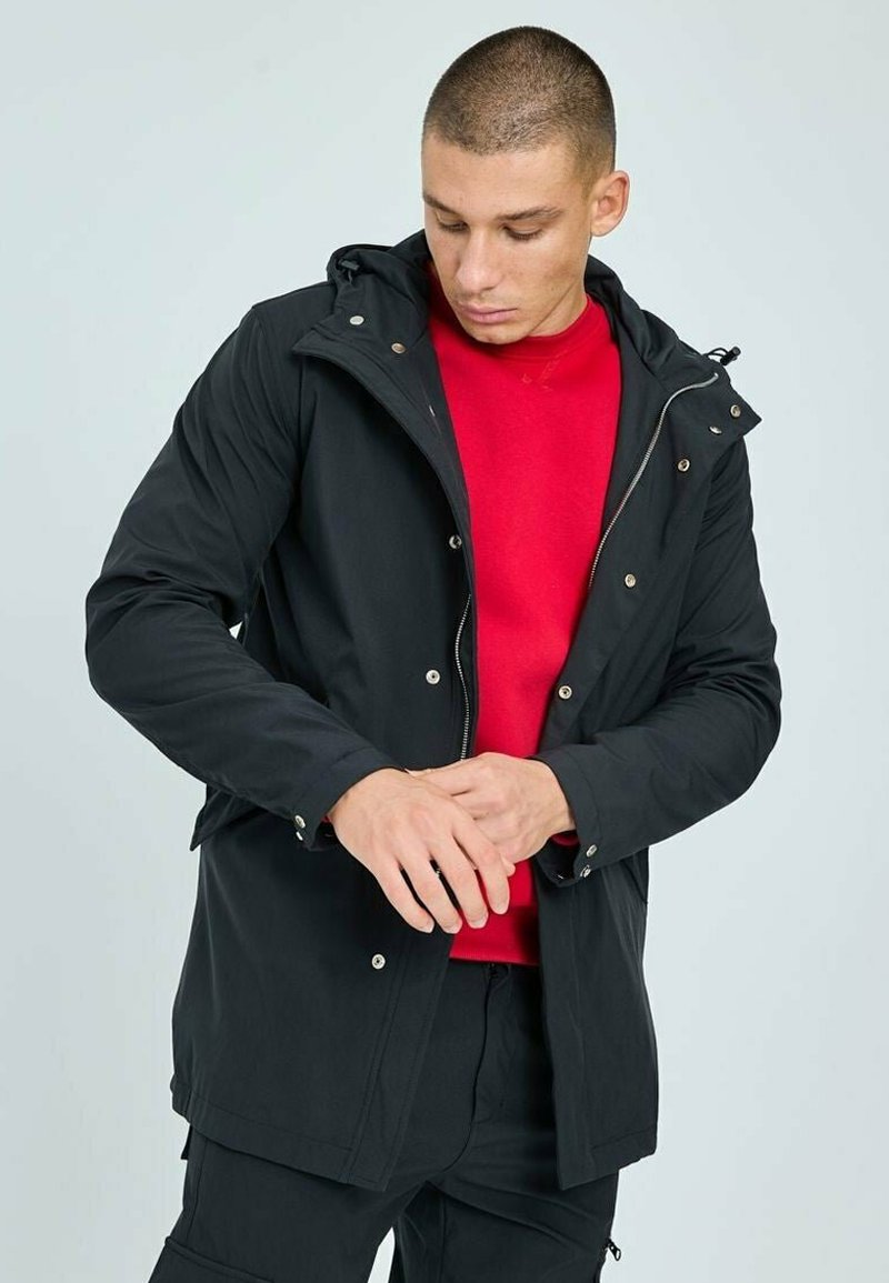 Veste extérieure noire avec fermeture éclair et boutons-pression, capuche ajustable et poches latérales, portée sur un pull à col rond rouge.