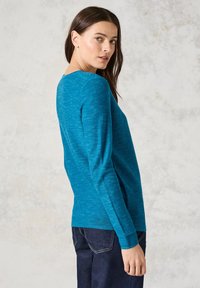 Türkiser Pullover mit langen Ärmeln und strukturiertem Rippenmuster sowie eng anliegenden Bündchen, kombiniert mit dunklen Jeans. Der Hintergrund ist neutral und strukturiert.