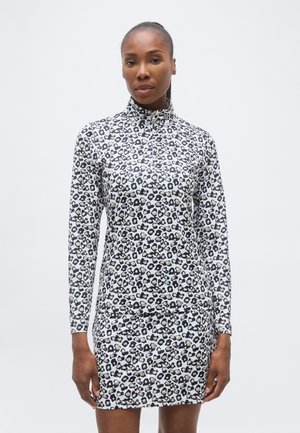 Vrouw die een witte leopardprint jurk met lange mouwen en een hoge kraag draagt, staand tegen een effen lichte achtergrond.