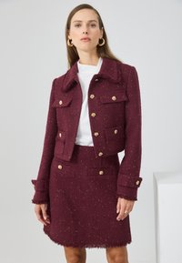 Bordeaux tweed sako s zlatými gombíkmi a dvoma prednými vreckami, v kombinácii s ladiacou sukňou so strapcovým spodným okrajom. Nosené na bielo.