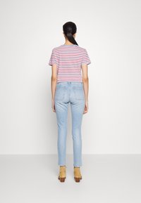 Woman with black ponytail wearing a red and white striped t-shirt, light blue high-waisted jeans, and tan ankle boots, standing facing away.