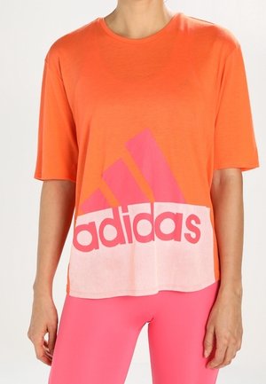 Osoba ubrana w pomarańczową koszulkę Adidas z dużym różowym logo i jasnoróżowe legginsy, stojąca na neutralnym tle.