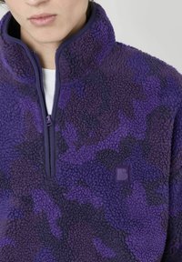 Pull en polaire violet avec col montant, fermeture éclair et surface douce et texturée. Présente un motif camouflage subtil et un patch logo sur la poitrine.