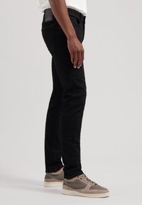 Jeans de denim negro con un corte ajustado, que presentan un parche de cuero en la cinturilla y detalles de costura. Combinados con zapatillas en gris claro.