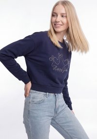 Fiatal szőke hajú nő sötétkék pulóvert visel, amelyen strasszköves korona díszeleg, és világoskék farmert, miközben mosolyogva oldalra néz.