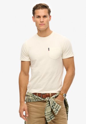 Lys beige kortærmet t-shirt med en brystlomme og mærkebadge. Sammen med khaki bukser og en grøn ternet skjorte bundet om livet.