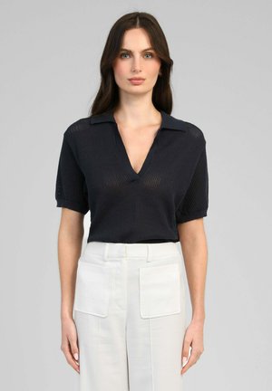 Femme aux longs cheveux bruns portant un polo en maille noire à manches courtes et un pantalon blanc taille haute avec des poches plaquées à l'avant.