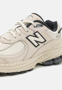 Beige sportcipő, textúrált hálós panelekben, fekete kiemelésekkel és egy jellegzetes fekete "N" logóval. Fűzőzáras és párnázott talppal rendelkezik.