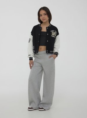 Ragazza adolescente in piedi con la mano in tasca indossa una giacca varsity bianca e nera, un top corto nero, pantaloni larghi grigi e scarpe da ginnastica su sfondo semplice.