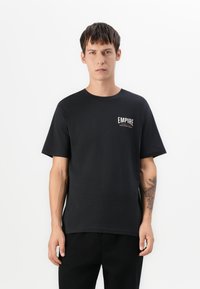 Ung mand med kort mørkt hår, iført en sort "Empire Fight Club" T-shirt og sorte bukser, stående imod en hvid baggrund.
