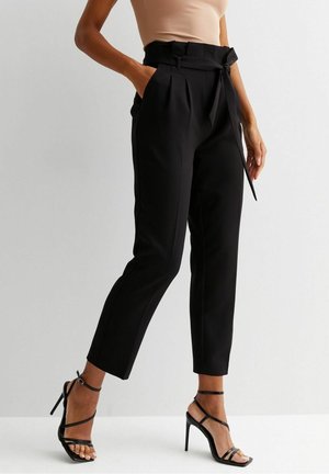 Vrouw draagt zwart hooggetailleerde taps toelopende broek met een gebonden riem en zwarte sandaal met hoge hak en bandjes, staande tegen een effen achtergrond.
