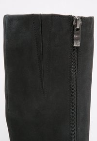 Botte noire en daim haute jusqu'au genou avec une texture lisse, dotée d'une fermeture éclair sur le côté et d'un détail de couture décoratif le long de la ligne de fermeture.