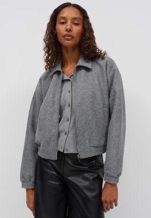 Vrouw die een grijze jas met ritssluiting draagt over een grijs overhemd met knopen, gecombineerd met zwarte leren broek, staand tegen een eenvoudige lichte achtergrond.