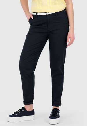 Vrouw draagt zwarte omslagbroek, wit gevlochten riem, geel topje en zwarte platformsneakers met witte zolen, staat met één hand in de zak.