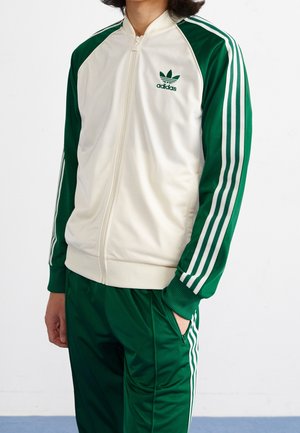 Persoon draagt een groen-wit Adidas trainingsjack met ritssluiting en bijpassende groene trainingsbroek met witte strepen.