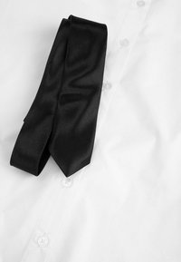 Czarny jedwabny krawat ułożony na zapiętej białej koszuli z trzema widocznymi guzikami na jednolitym tle.