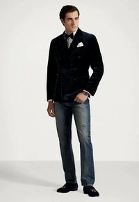 Tmavě modrý sametový dvouřadý blejzr přes bílou košili s motýlkem, doplněný o džíny s viditelným opotřebením a tmavé mokasíny.