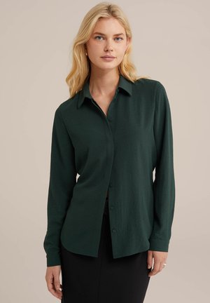 Dark green button-up shirt with long sleeves, a collar, and a soft texture. Features a relaxed fit and straight hem, worn over a black skirt.