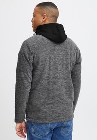 Grå fleecejakke med sort hætte, der har en tekstureret overflade og lynlåslommer, båret over blå denimjeans.