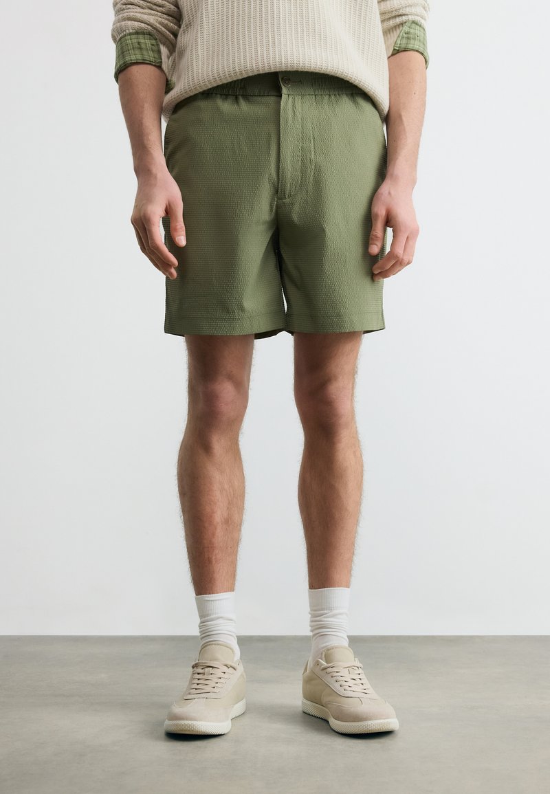 Άνδρας που φοράει χακί υφασμάτινες βερμούδες, μπεζ αθλητικά παπούτσια, λευκές κάλτσες και κρεμ πουλόβερ με γυρισμένα πράσινα μανίκια, που στέκεται σε γκρι δάπεδο.