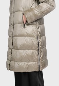 Ljus taupefärgad vadderad jacka med quiltad design, utrustad med sidofickor med dragkedja och en slät, glänsande yta. Halvlång med skräddarsydd passform.