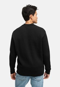 Mand med ryggen til iført en sort langærmet sweater og lyseblå jeans foran en ensfarvet lys baggrund.