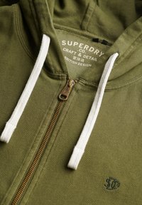 Sudadera con cremallera en verde oliva con cordones blancos, que presenta una cremallera de metal y un sutil logotipo bordado en el frontal. Textura suave, diseño casual.