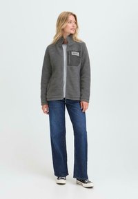 Kvinne iført grå fleecejakke med brystlomme, blå jeans og svarte joggesko, stående og ser til siden på en ensfarget bakgrunn.