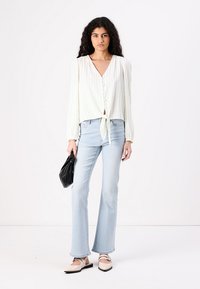 White vertically striped blouse with a front tie, paired with light blue flared jeans. Holds a black bag; wears light-colored shoes.