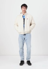 Lys beige pufferjakke med ribbet krave, lynlåslukning og forlommer. Bæres over en mørkeblå skjorte og lyse jeans med sorte sneakers.