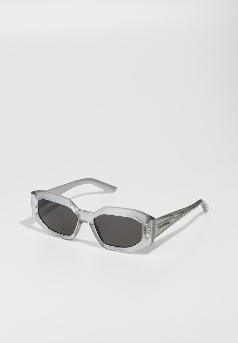 Lunettes de soleil octogonales grises avec des verres sombres. Monture grise transparente au toucher lisse et branches fines et élégantes. Branding discret sur le côté.
