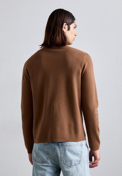 Mann mit schulterlangem Haar, der einen braunen Langarmpullover und helle blaue Jeans trägt und von einem schlichten Hintergrund abgewandt steht.