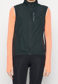 Black lightweight zip-up vest with high collar, elastic hem, and white logo. Worn over a long-sleeve, bright orange athletic top.