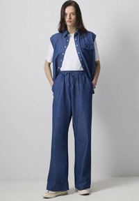 Denim outfit featuring a sleeveless blue shirt with pockets and matching loose-fitting blue pants, styled with a white t-shirt underneath.